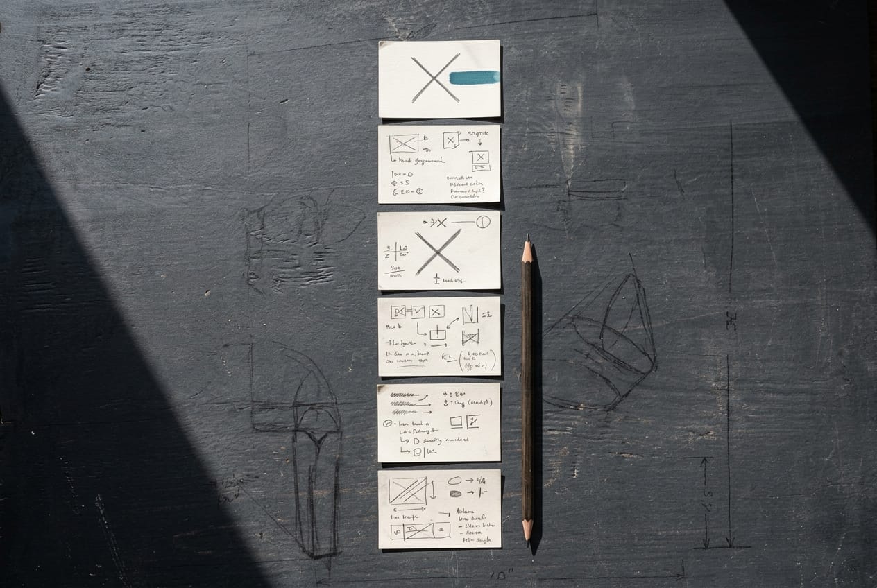 Huit fiches cartonnées disposées en colonne sur un bureau, chacune annotée à la main, deux d'entre elles portant une croix au crayon rouge